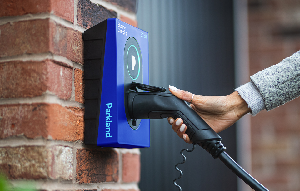 A person's hand grasping the handle of an electric vehicle charger. The charger is Parkland blue with the Parkland logo on the side